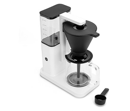 Eine weiße Kaffeemaschine mit Glaskanne und schwarzem Filter ist zusammen mit einem schwarzen Messlöffel abgebildet.