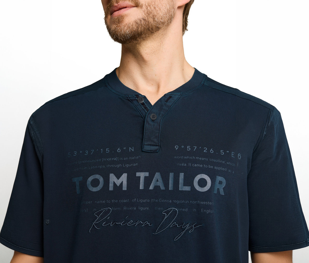 Mann trägt ein dunkelblaues Henley-T-Shirt mit dem Aufdruck 'TOM TAILOR', 'Riviera Days' und geografischen Koordinaten.