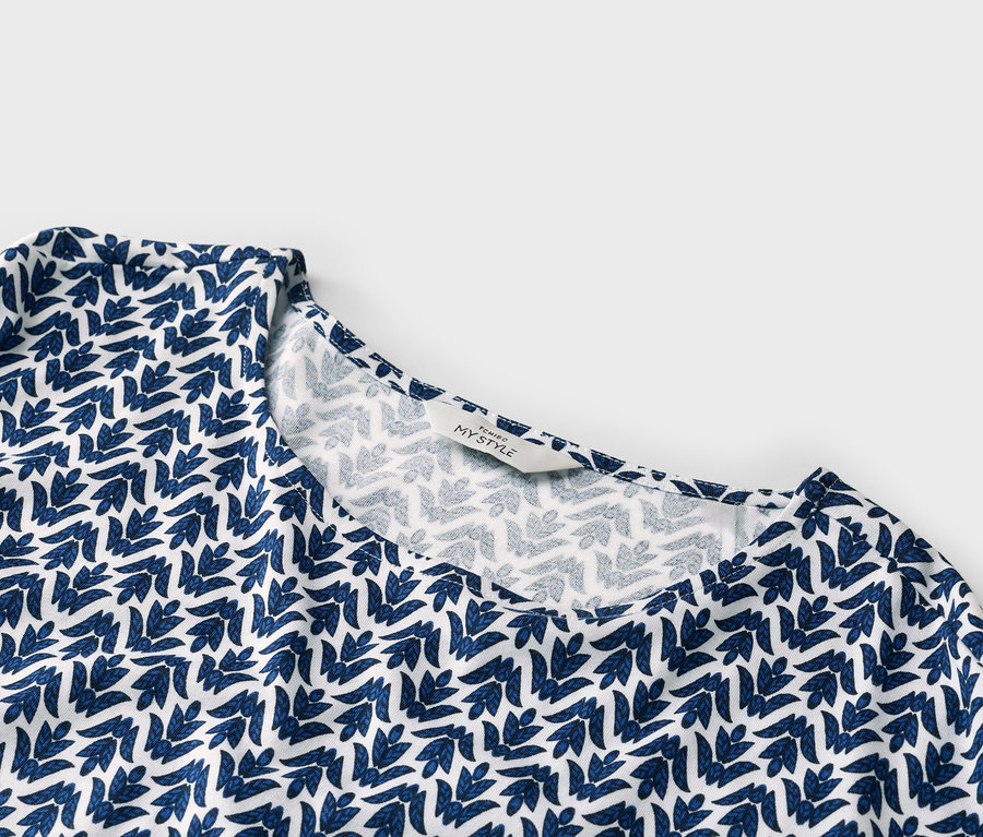 Nahaufnahme eines weissgrundigen Oberteils mit blauem Blattmuster und einem Etikett mit der Aufschrift "TCHIBO MY STYLE".