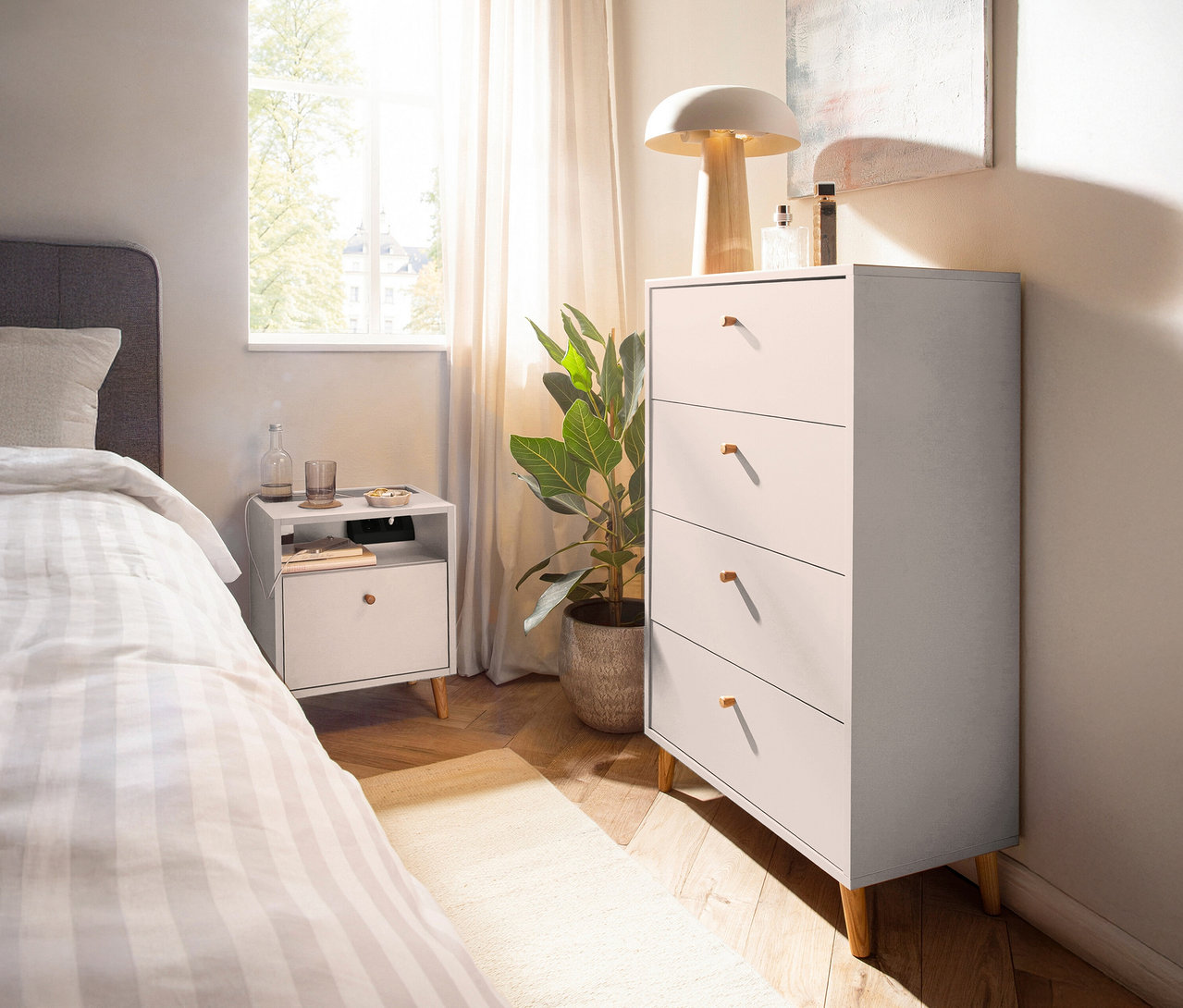 Schlafzimmer mit Kommode »Bjarne« in mattem Weiß, Nachttisch, Bett und Zimmerpflanze.
