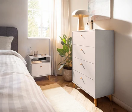 Schlafzimmer mit Kommode »Bjarne« in mattem Weiß, Nachttisch, Bett und Zimmerpflanze.