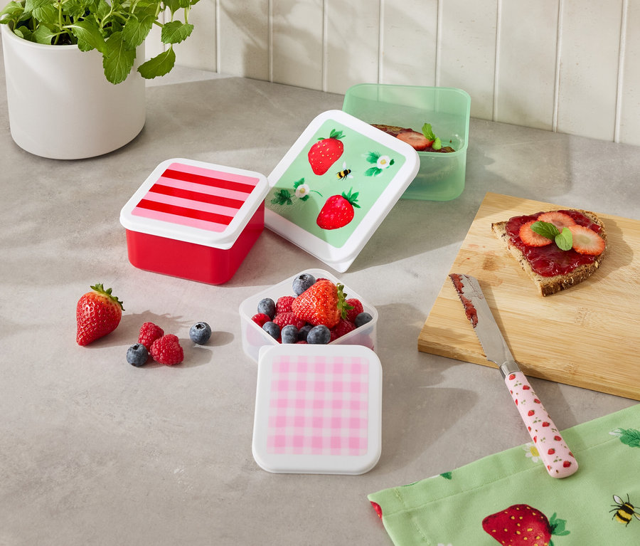 Mehrere bunte Frischhaltedosen stehen auf einer Küchentheke, einige gefüllt mit Früchten, neben einem belegten Brot und einem Messer.