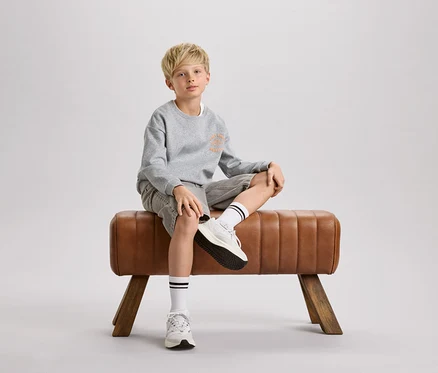 Ein blonder Junge sitzt auf einem braunen Turnbock. Er trägt ein graues Sweatshirt, Shorts und weiße Socken mit schwarzen Streifen.