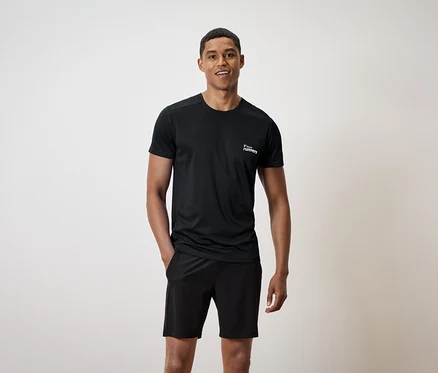Mann mit schwarzem T-Shirt mit Aufdruck und schwarzen Shorts, eine Hand in der Hosentasche.