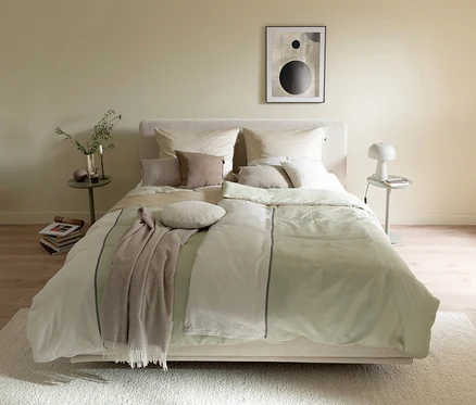 Ein Schlafzimmer mit einem Bett, das mit SCHÖNER WOHNEN-Kollektion Bettwäsche »PLACES« in Hellgrün und Beige mit grauem Streifen bezogen ist. Auf dem Bett liegen verschiedene Kissen und eine braune Decke. Links steht ein Beistelltisch mit einer Vase, rechts ein Beistelltisch mit einer weißen Lampe. Über dem Bett hängt ein abstraktes Bild.