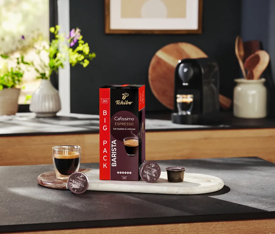 Ein Glas Espresso steht neben einer Packung Tchibo Cafissimo Barista Kapseln auf einem Küchentisch.
