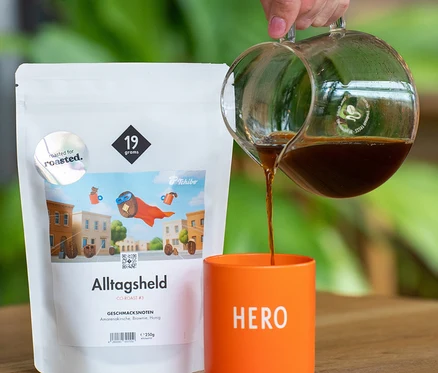 Eine Hand gießt Kaffee aus einer Glaskaraffe in einen orangefarbenen Becher mit „HERO“-Aufschrift. Daneben steht eine weiße 250g-Packung 19grams x roasted. - Alltagsheld Co-Roast #3 Omni-Roast Kaffeebohnen.