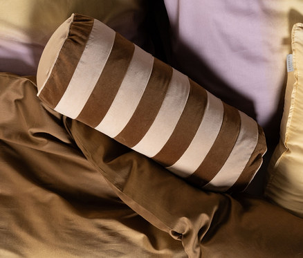 Bett mit Ombre-Bettwäsche von Gelb zu Lila, teilweise bedeckt von einer braunen Decke. Darauf liegen ein hellgelbes Kissen, ein braun-beige gestreiftes Nackenkissen und ein rundes braunes Essenza Samt-Dekokissen »Bell« mit Füllung.