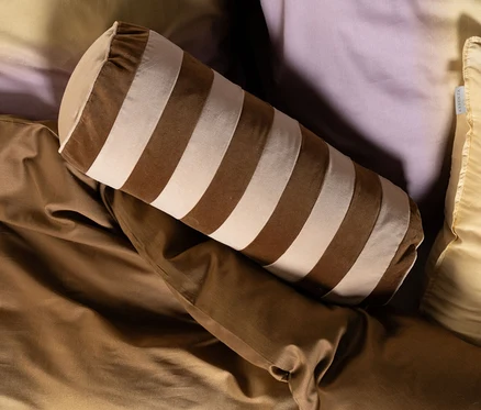 Bett mit Ombre-Bettwäsche von Gelb zu Lila, teilweise bedeckt von einer braunen Decke. Darauf liegen ein hellgelbes Kissen, ein braun-beige gestreiftes Nackenkissen und ein rundes braunes Essenza Samt-Dekokissen »Bell« mit Füllung.