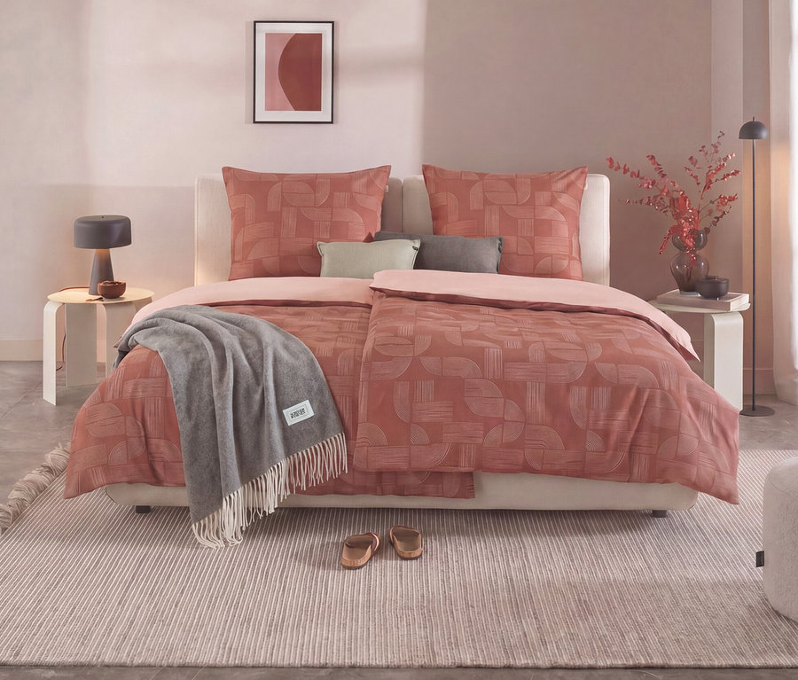 Ein Schlafzimmer mit einem Bett, das mit der SCHÖNER WOHNEN – Kollektion Bettwäsche in Normalgröße bezogen ist. Die Bettwäsche ist terrakottafarben mit einem geometrischen Muster und einer hellrosa Unterseite. Ein graues Plaid liegt auf dem Bett. Links und rechts stehen Nachttische mit Lampen, rechts ein Hocker mit Teekanne und Tasse.