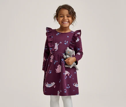 Ein lächelndes kleines Mädchen in einem lila Kleid mit Rüschen und Katzenmuster, das ein graues Plüschkätzchen hält.