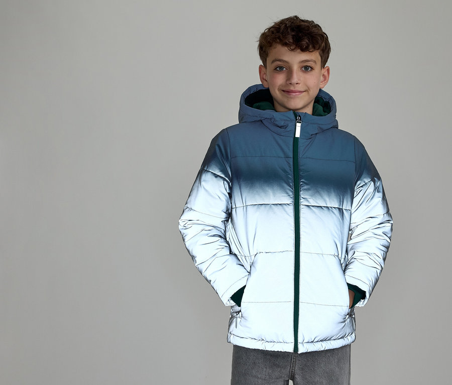 Ein lächelnder Junge mit lockigem Haar trägt eine Kapuzen-Steppjacke. Die Jacke hat einen Farbverlauf von Dunkelblau oben zu reflektierendem Silber unten und einen grünen Reißverschluss. Seine Hände stecken in den Jackentaschen.