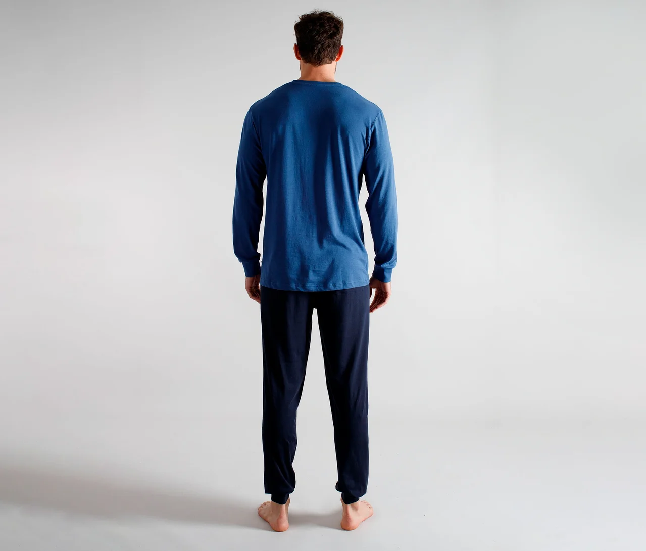 Mann von hinten in einem blauen Langarmshirt und einer dunkelblauen Hose, barfuß stehend.