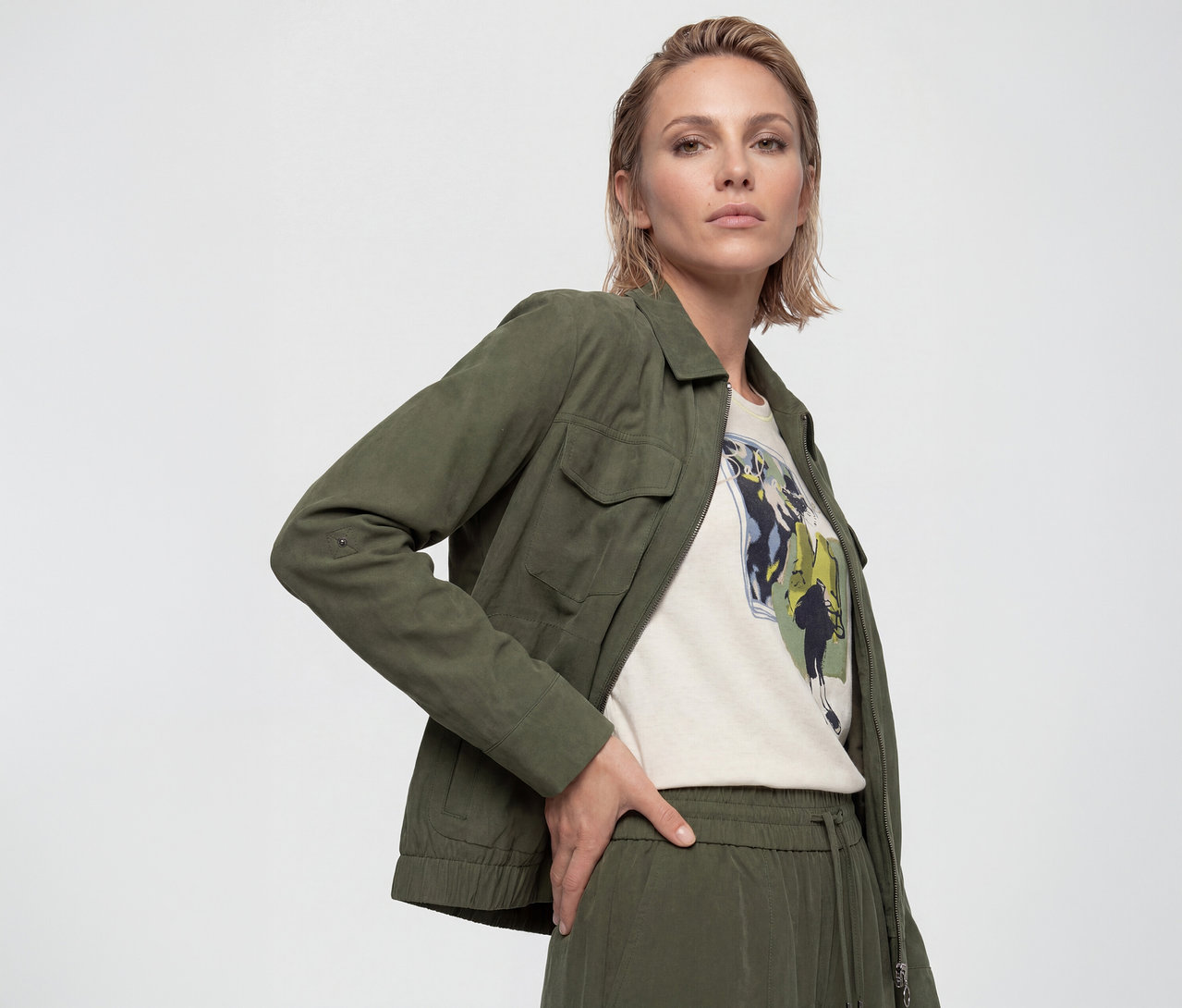 Frau mit kurzen blonden Haaren trägt eine grüne Utility-Jacke, ein helles T-Shirt mit Grafikprint und eine grüne Hose mit Kordelzug, posiert mit der Hand in der Hüfte.