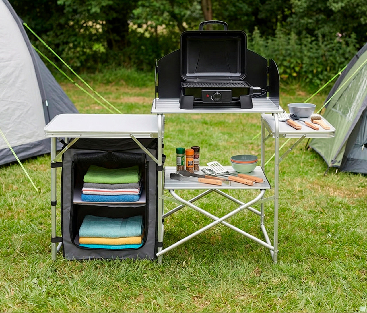 Faltbare Campingküche »Redwood S« auf grünem Gras vor zwei Zelten. Oben Elektrogrill, unten Gewürze, Kochutensilien, Schüsseln. Stofffach mit gefalteten Handtüchern.