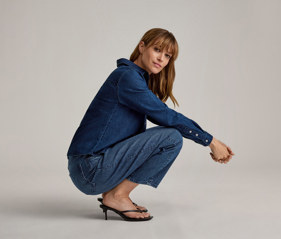 Eine Frau hockt, trägt ein dunkelblaues Jeanshemd, hellblaue Jeans und schwarze hochhackige Zehentrenner.