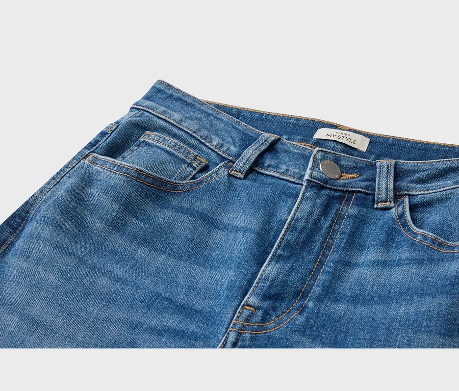 Nahaufnahme einer blauen Jeans, die die Vordertaschen, einen silbernen Knopf und ein weißes "TCHIBO MY STYLE"-Etikett am Bund zeigt.
