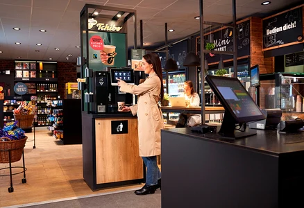 Eine Frau im beigen Trenchcoat bedient eine Tchibo Selbstbedienungs-Kaffeemaschine und hält einen Becher. Im Hintergrund arbeitet eine andere Frau an einem Laptop an einem Tresen im Minimarkt.