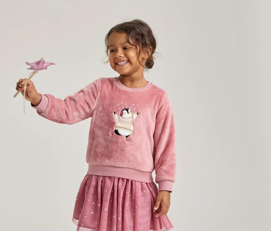 Lächelndes Mädchen in einem flauschigen rosa Pullover mit Pinguin-Motiv und einem rosa Rock mit Sternen, das einen rosa Sternenzauberstab hält.