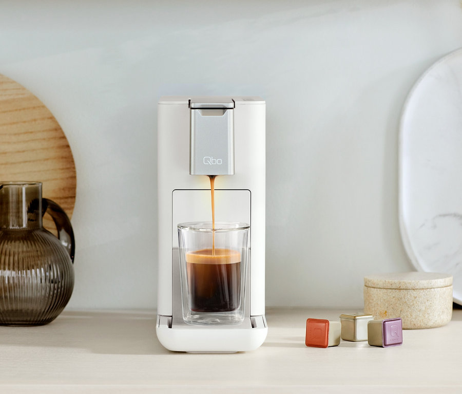 Qbo ICONIC Dark Chrome und Qbo ESSENTIAL Bright Sand Kaffeemaschine füllt ein Glas mit Kaffee. Daneben stehen Kapseln.