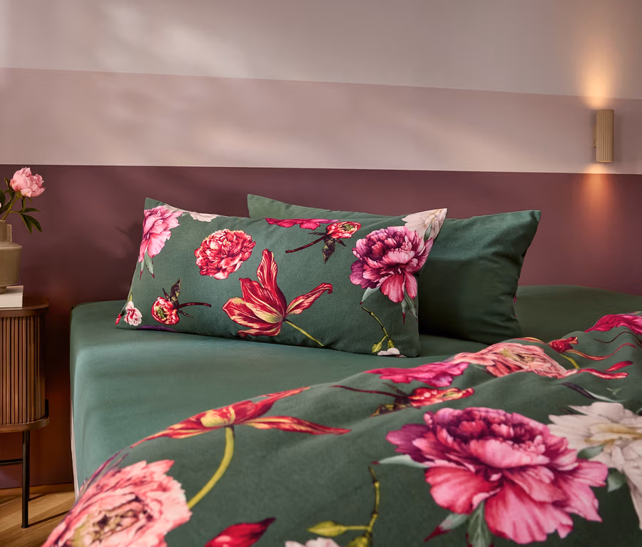 Ein Bett mit dunkelgrüner Bettwäsche, die mit großen rosa und roten Blumen gemustert ist, und einem einfarbigen grünen Kissen. Links steht ein Nachttisch mit einer Vase voller rosa Pfingstrosen. Die Wand ist zweifarbig in Rosa und Dunkelrot gestrichen.