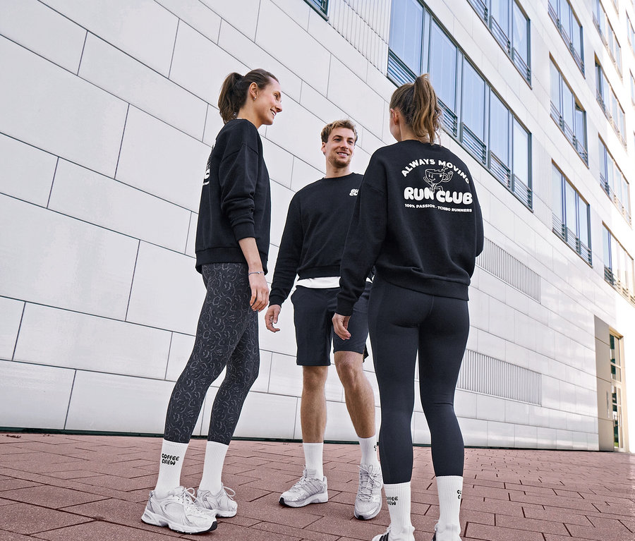 Drei Personen in schwarzen Sweatshirts und Sportkleidung unterhalten sich. Eine Frau trägt grau-schwarze Sportleggings »ActivePower« und Unisex Coffee Crew Tennissocken. Ein Mann trägt schwarze Sportshorts und weiße Socken. Eine andere Frau trägt schwarze Sportleggings »ActivePower« und ein Sweatshirt mit „RUN CLUB“-Logo.