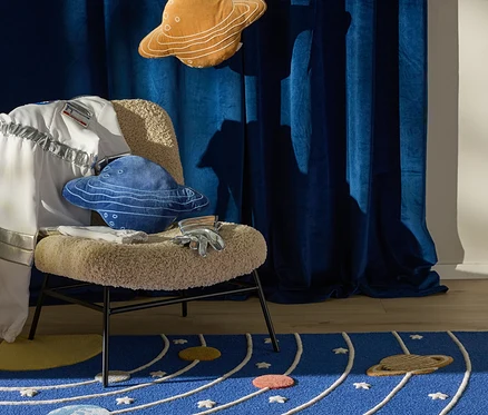 Ein blauer Teppich mit Sonnensystem-Design, Planeten und Umlaufbahnen. Ein Stuhl mit flauschigem beigem Sitz hält einen Astronautenanzugärmel, blaue und orange Planetenkissen sowie silberne Handschuhe. Blaue Samtvorhänge hängen im Hintergrund mit einer Raketen- und Planetendekoration.
