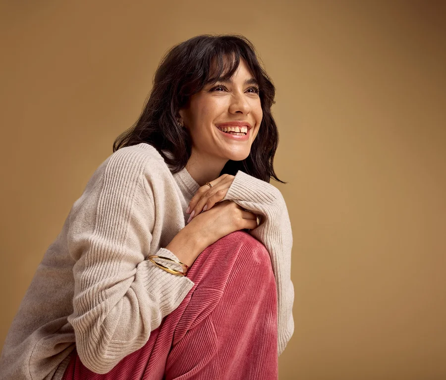 Lächelnde Frau mit dunklen Haaren, die einen hellbeigen Pullover und eine rosa Cordhose trägt, sitzt mit über den Knien verschränkten Händen. Sie trägt ein goldenes Armband und einen Ring.