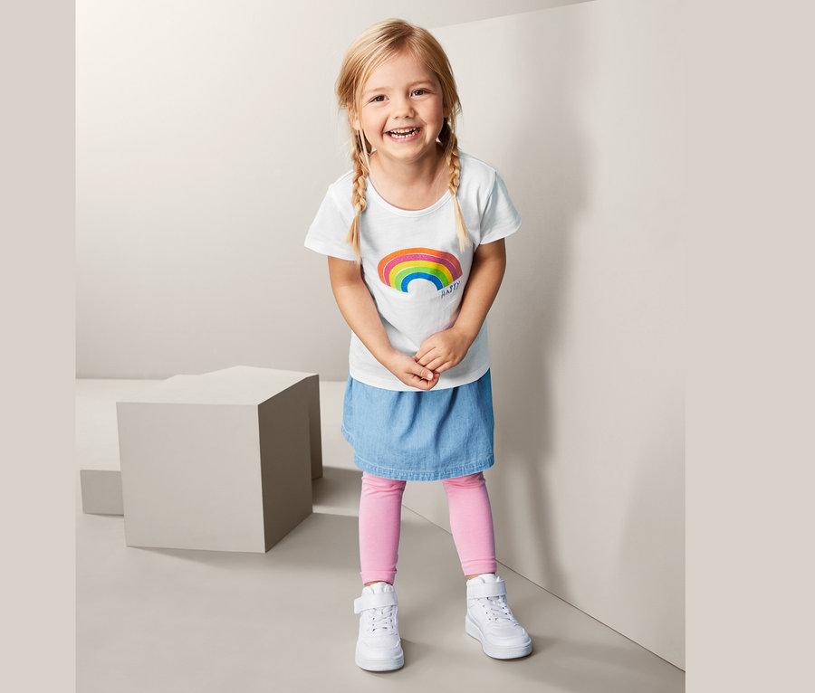 Lächelndes Mädchen trägt ein weißes T-Shirt mit Regenbogen, einen Jeansrock und rosa Leggings.
