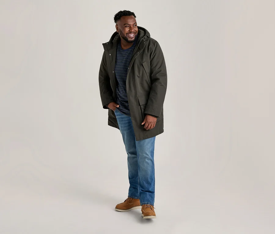 Lächelnder Mann in einem dunkelgrünen Parka, gestreiftem T-Shirt, blauen Jeans und braunen Schuhen, steht mit einer Hand in der Tasche.