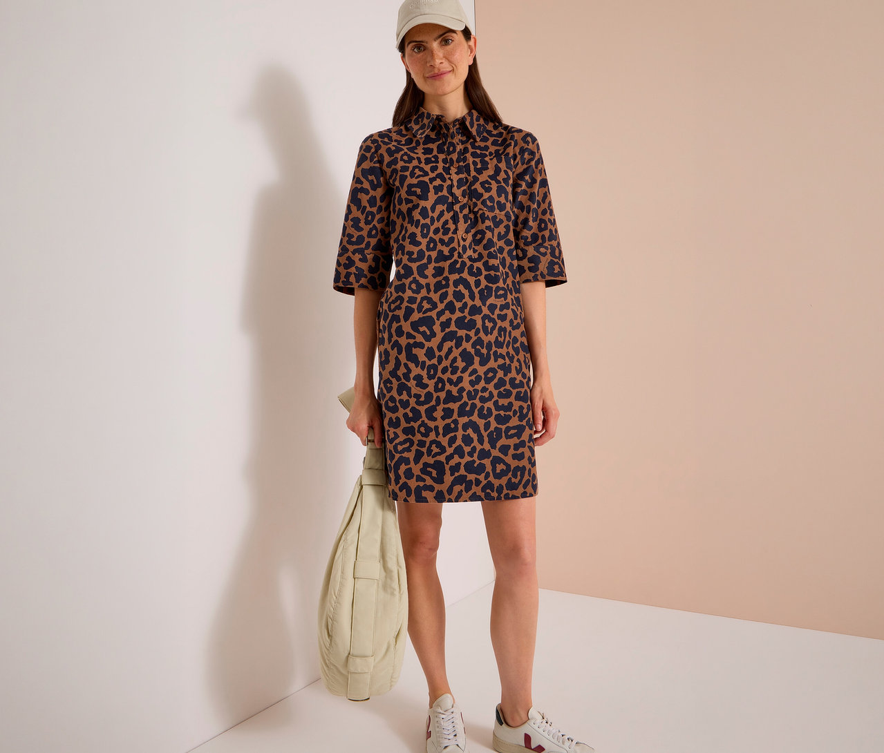 Frau trägt ein CECIL Cordkleid mit braunem und schwarzem Leopardenmuster, eine beige Baseballkappe und weiße Sneaker, eine hellgrüne Puffer-Tasche haltend.