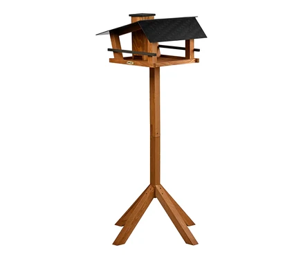 Ein hölzernes Vogelhaus mit schwarzem Dach steht auf einem hohen Holzpfahl mit Dreibein-Standfuß.