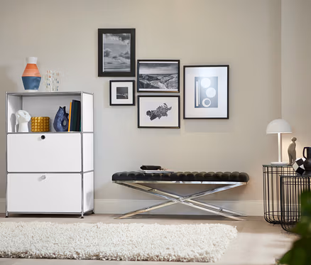 Ein Wohnzimmer zeigt ein Metallregal mit 2 weißen Klappenfächern, eine Design-Bank mit Metallgestell und einen Design-Barwagen aus Metall.