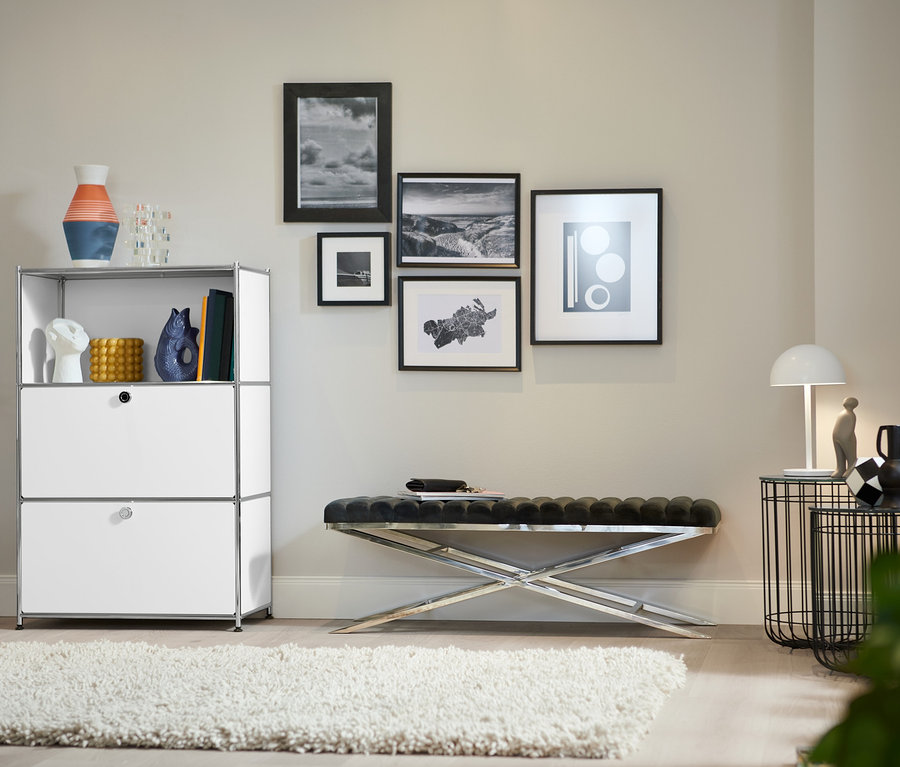 Ein Wohnzimmer zeigt ein Metallregal mit 2 weißen Klappenfächern, eine Design-Bank mit Metallgestell und einen Design-Barwagen aus Metall.
