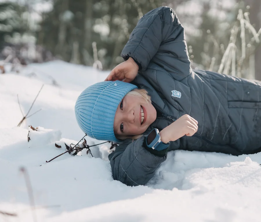 Ein lächelndes Kind liegt im Schnee, trägt eine graue Winterjacke, eine hellblaue Mütze und eine blaue Kids Watch Xplora X6 Play am Handgelenk.