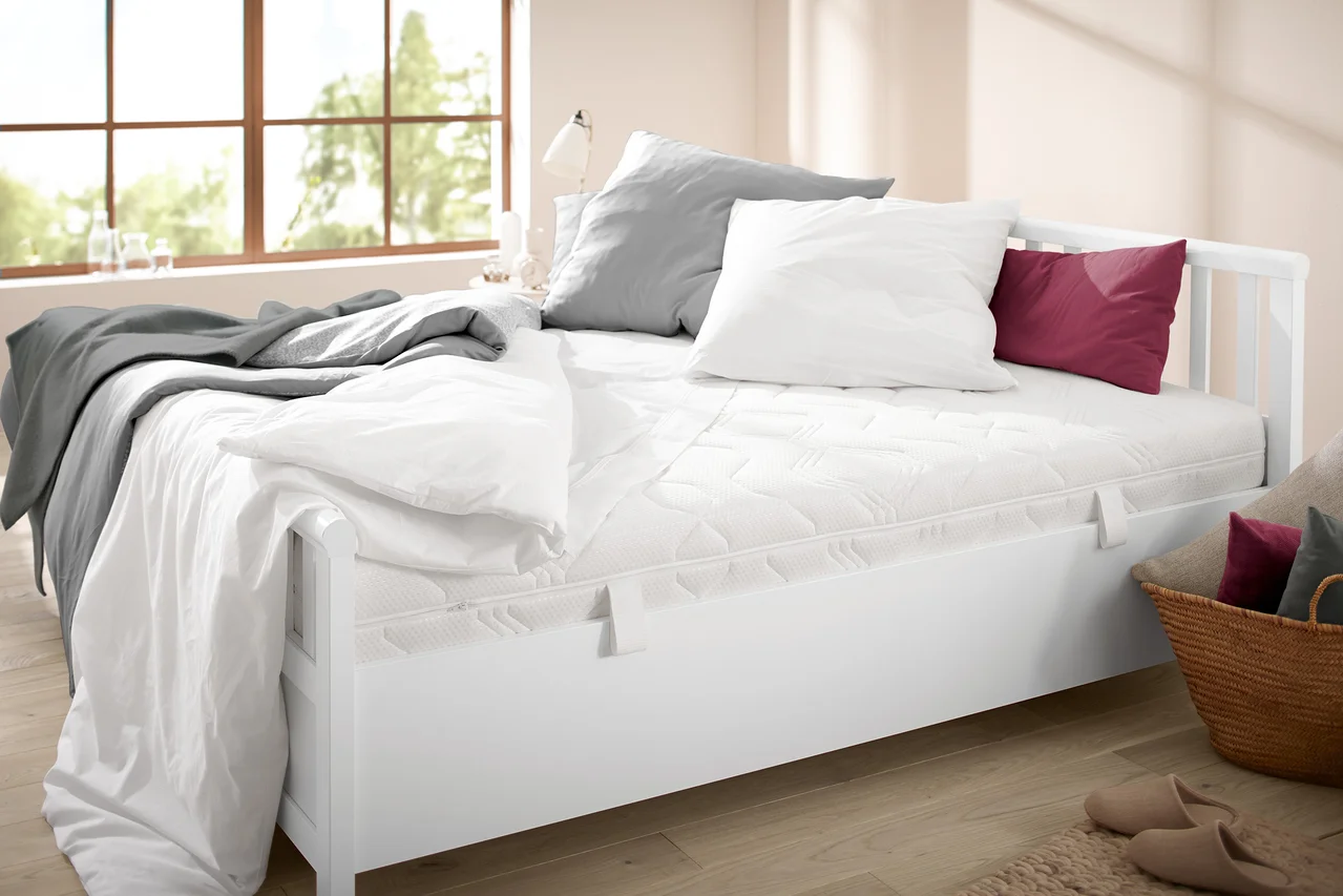 Bett mit weißem Rahmen und gesteppter Matratze. Auf dem Bett liegen eine graue Decke, ein weißes und ein graues Kissen sowie ein weinrotes Kissen.
