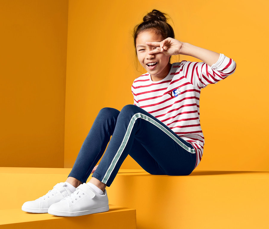 Ein Mädchen sitzt vor einem gelben Hintergrund und bedeckt ein Auge. Sie trägt 2 Leggings und ein gestreiftes Shirt.