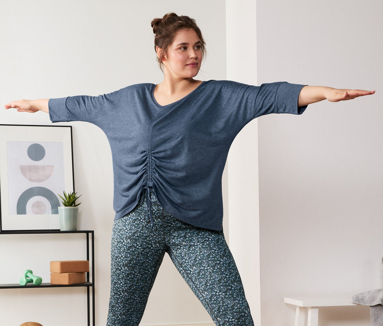 Eine Frau macht Yoga auf einer Matte, sie trägt eine blaue Wende-Sporttight und ein blau meliertes 3/4-Sport-und-Yogashirt.