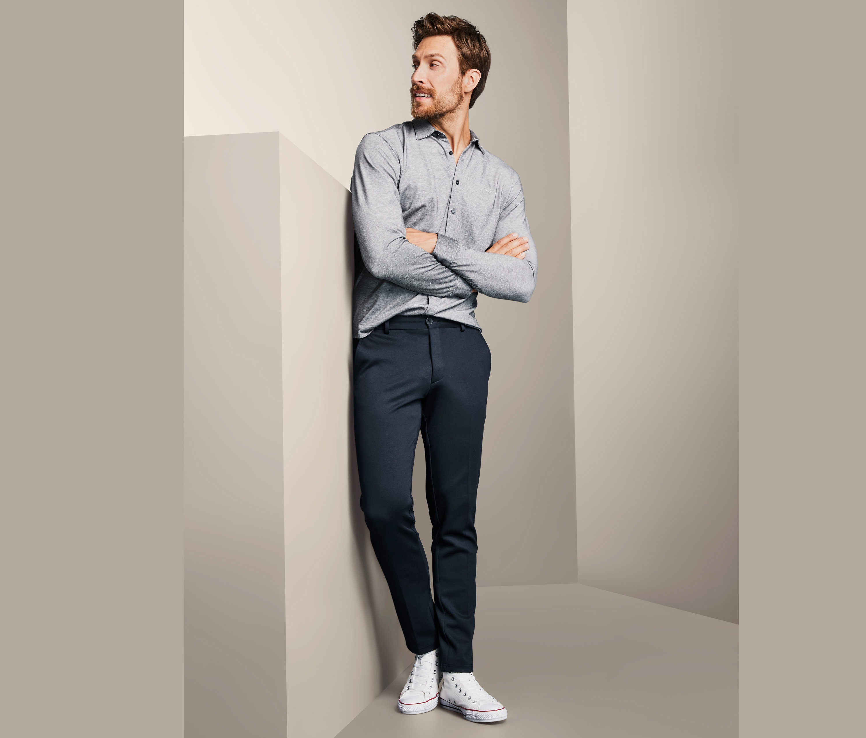 Mann lehnt an Wand, trägt graues Hemd und Jersey-Chino.
