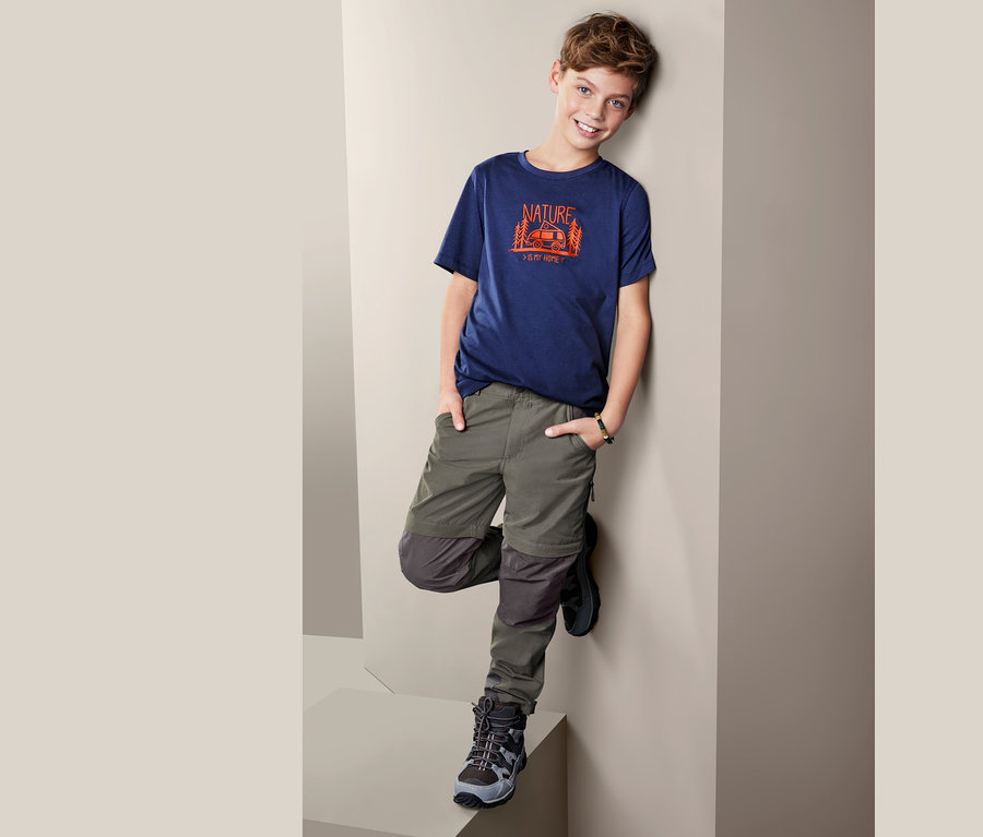Junge posiert an eine Wand gelehnt in einem T-Shirt und einer 2-in-1-Funktionshose.