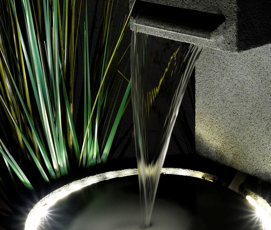 Wasser fließt aus einem steinernen Brunnen in eine beleuchtete Schale, neben der grüne Pflanzen wachsen. Dobar Gartenbrunnen mit Schale.