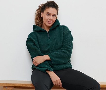 Frau sitzt auf Bank, trägt Bodyforming-Sporthose und dunkelgrünen Teddy-Kapuzenpullover.