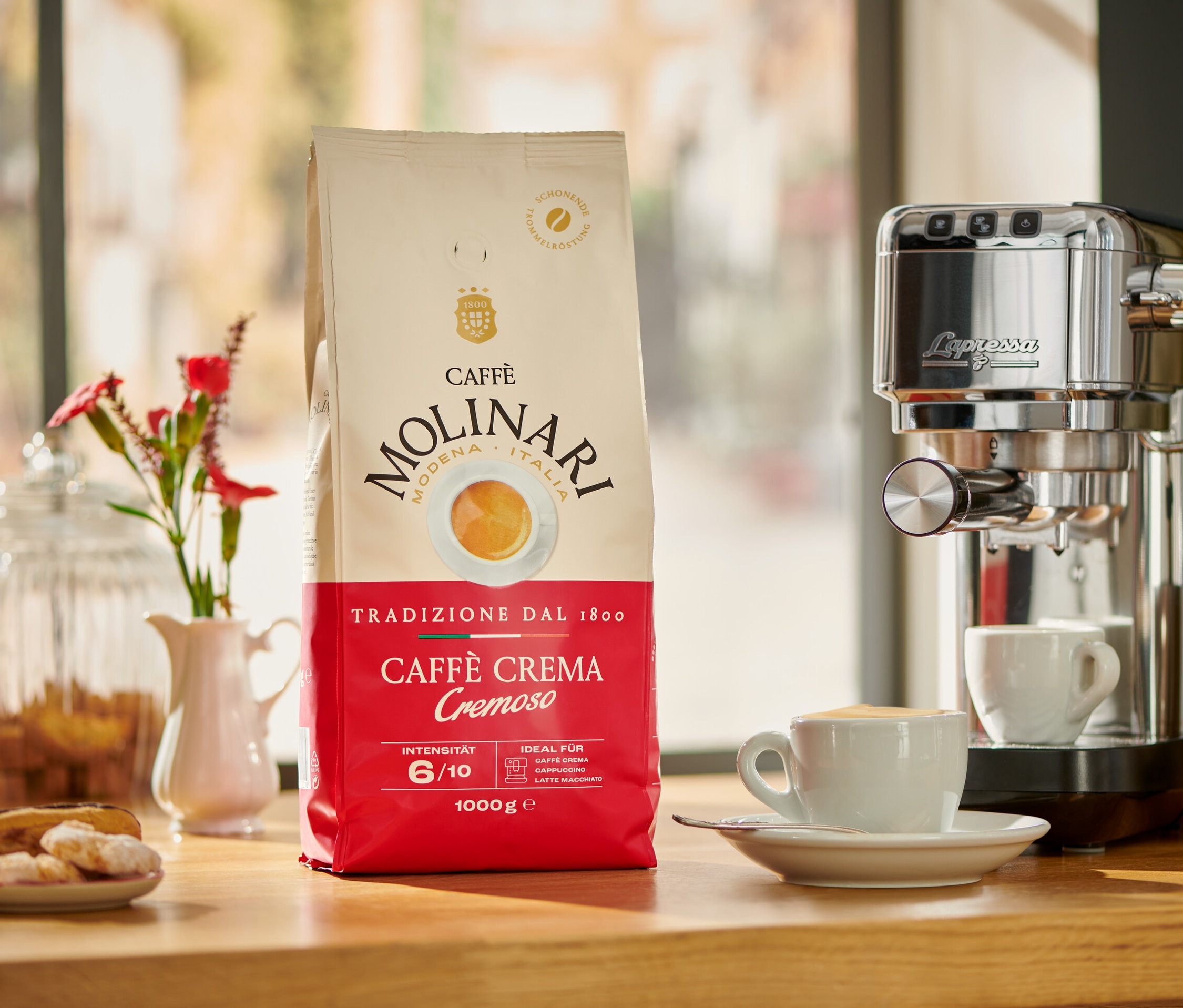 Eine Packung Caffè Molinari Caffè Crema Cremoso neben einer Kaffeemaschine und einer Tasse Kaffee.