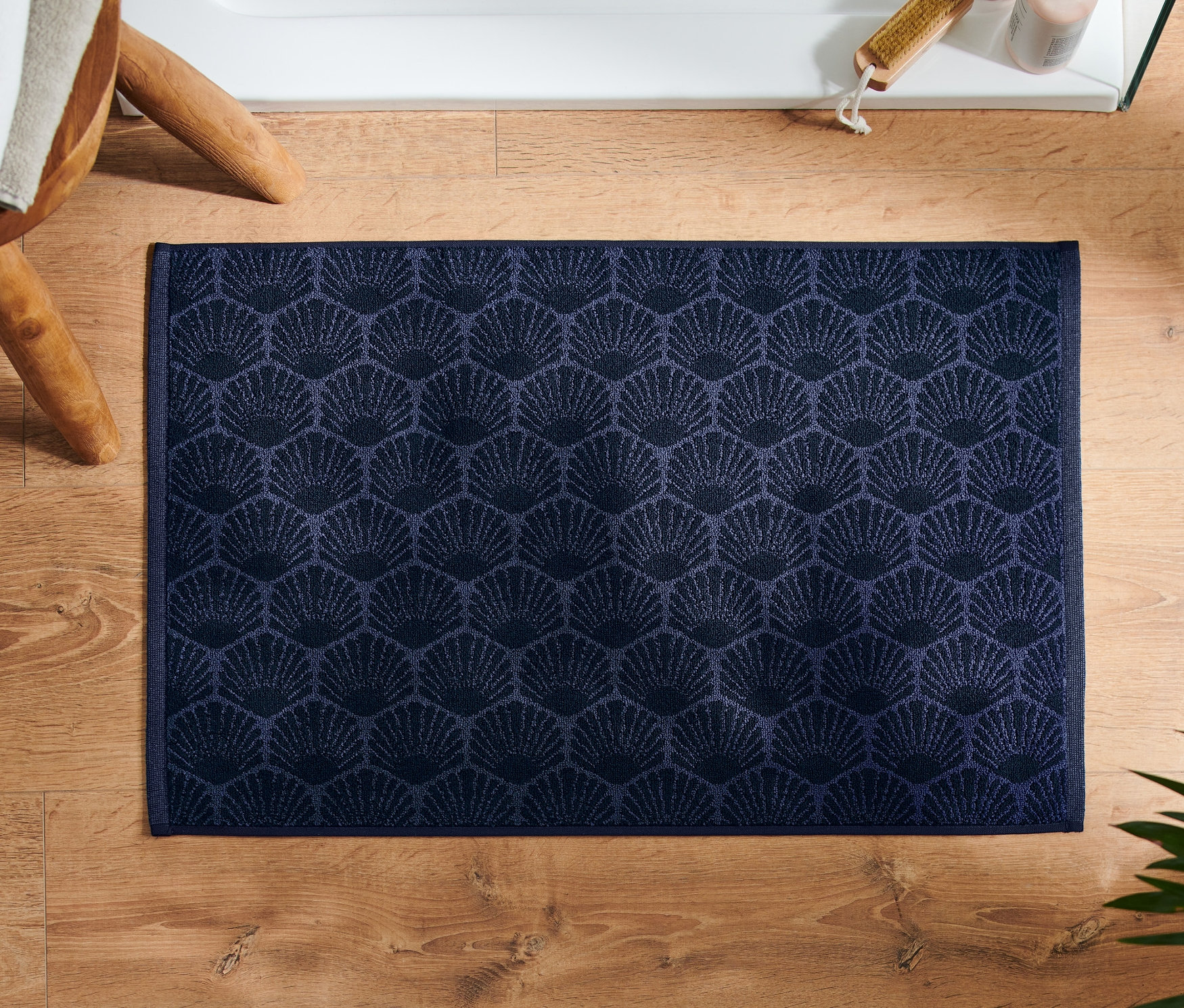 Marineblauer Duschvorleger »Muschel« liegt auf Holzboden neben Hocker und Pflanze.
