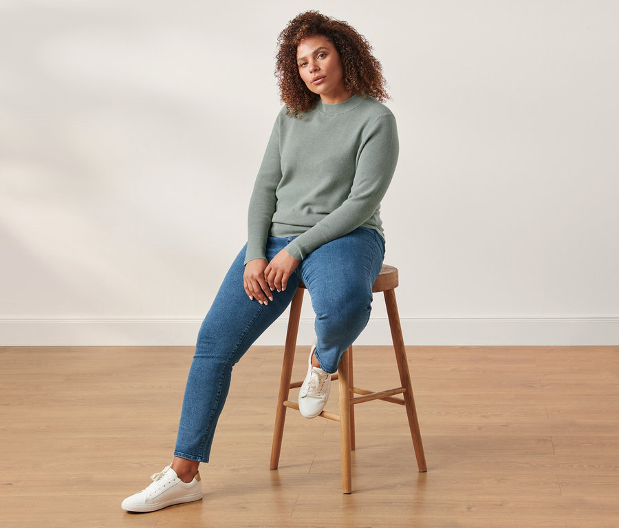Frau sitzt auf einem Holzhocker und trägt einen Strickpullover und eine Slimfit-Jeans.