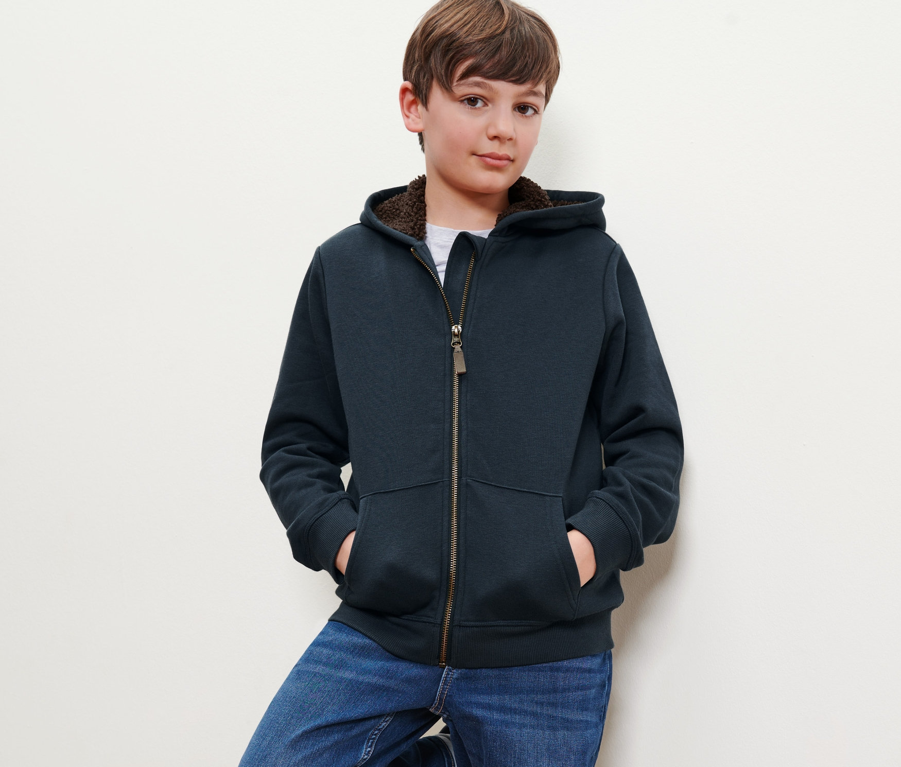 Ein Junge posiert in einer Kinder-Sweat-Jacke mit Kapuze, die Hände in den Taschen, an eine Wand gelehnt.