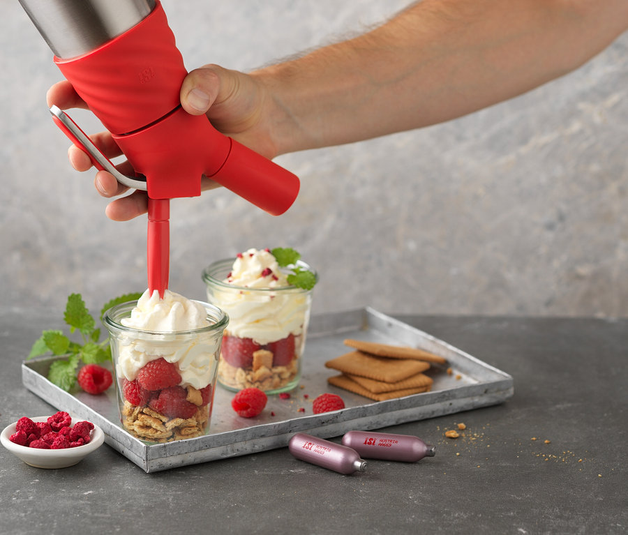 Eine Hand füllt ein Glas mit Himbeeren und Keksen mit Schlagsahne mit einem roten ISI Creative Whip 0,5 l. Auf einem Tablett befinden sich weitere Gläser, Kekse und Patronen.