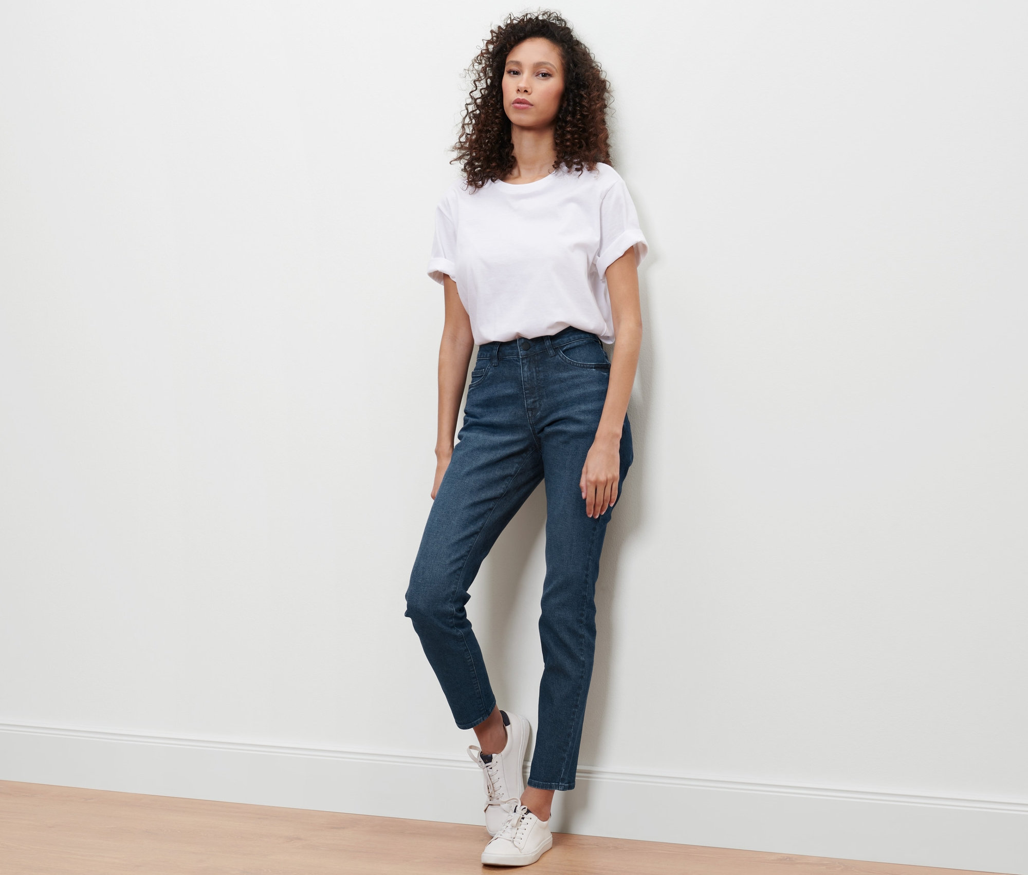 Eine Frau lehnt an einer Wand und trägt eine Slim-Denim »Fit Emma« in Dunkelblau.