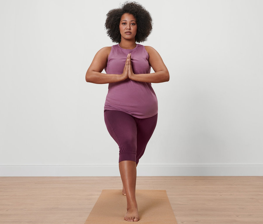 Frau in 3/4-Yogahose in Mauve und Sporttop in Mauve steht auf einer Yogamatte.