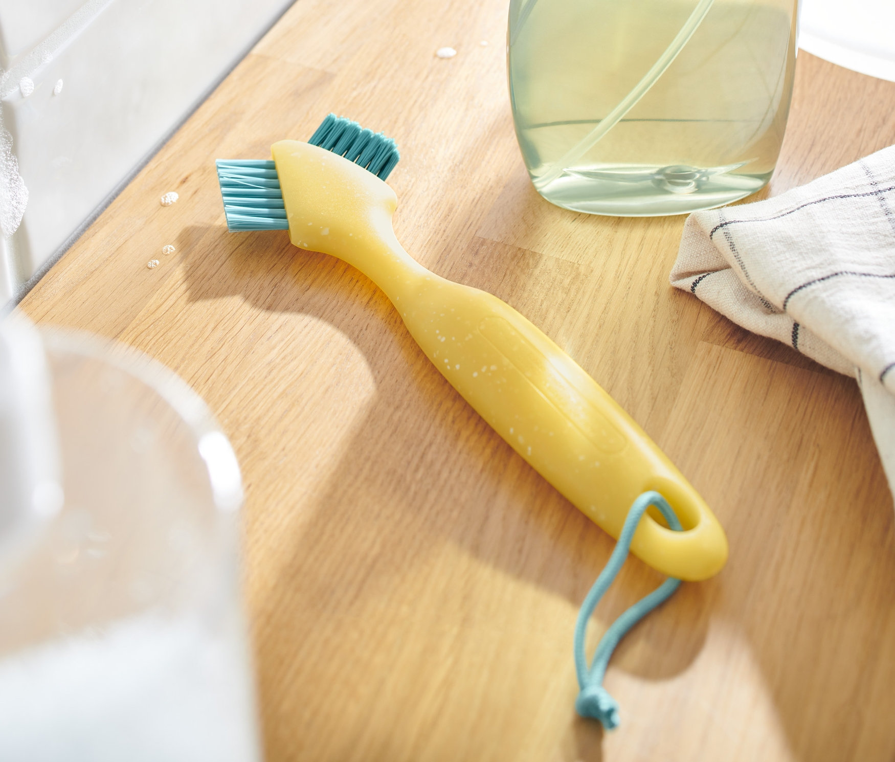 Gelbe Fugenbürste mit blauen Borsten liegt auf einer Holzoberfläche neben Reinigungsmittel und Tuch.